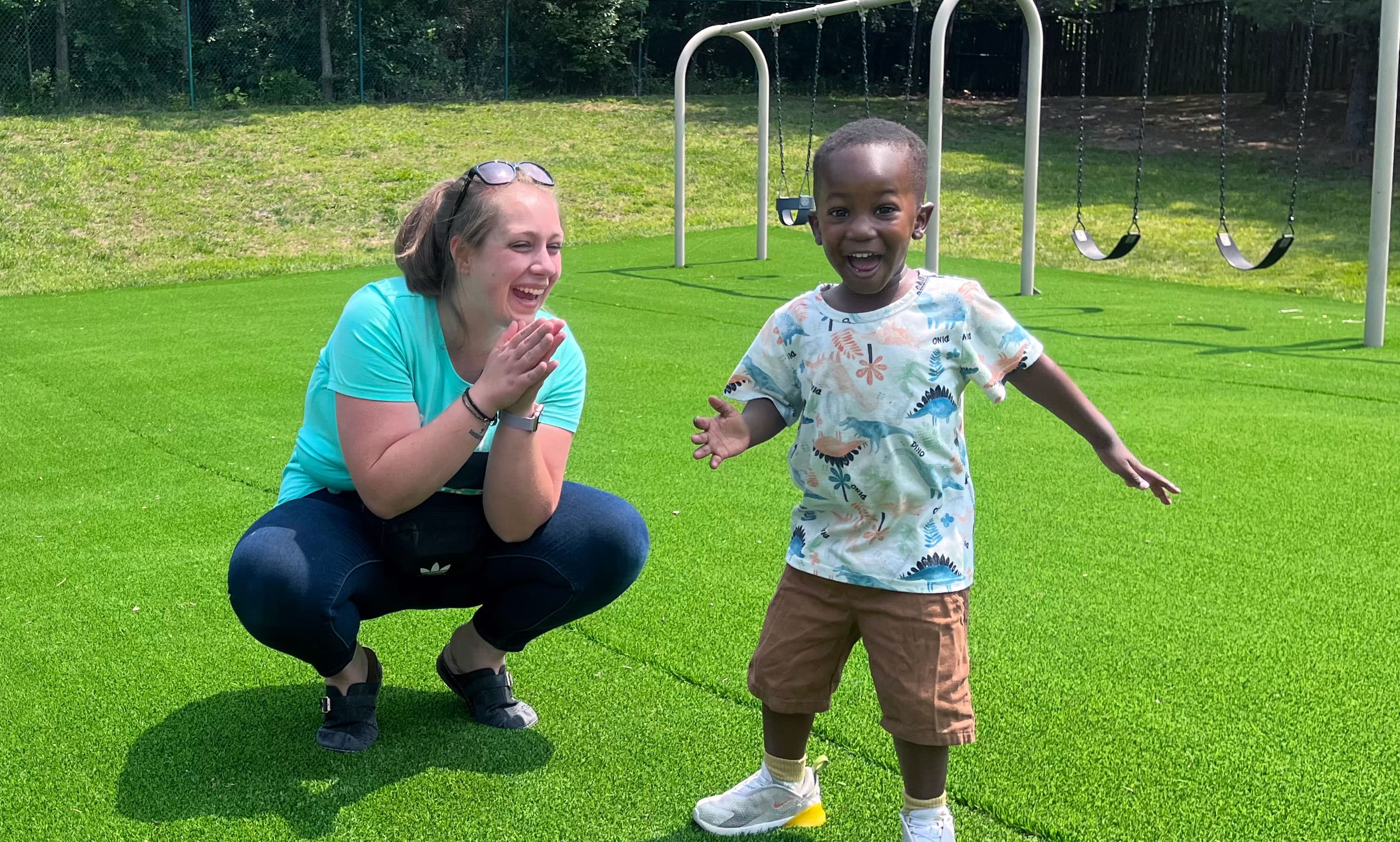 A teacher crouching and clapping with delight as a smiling toddler runs toward her on the playground turf