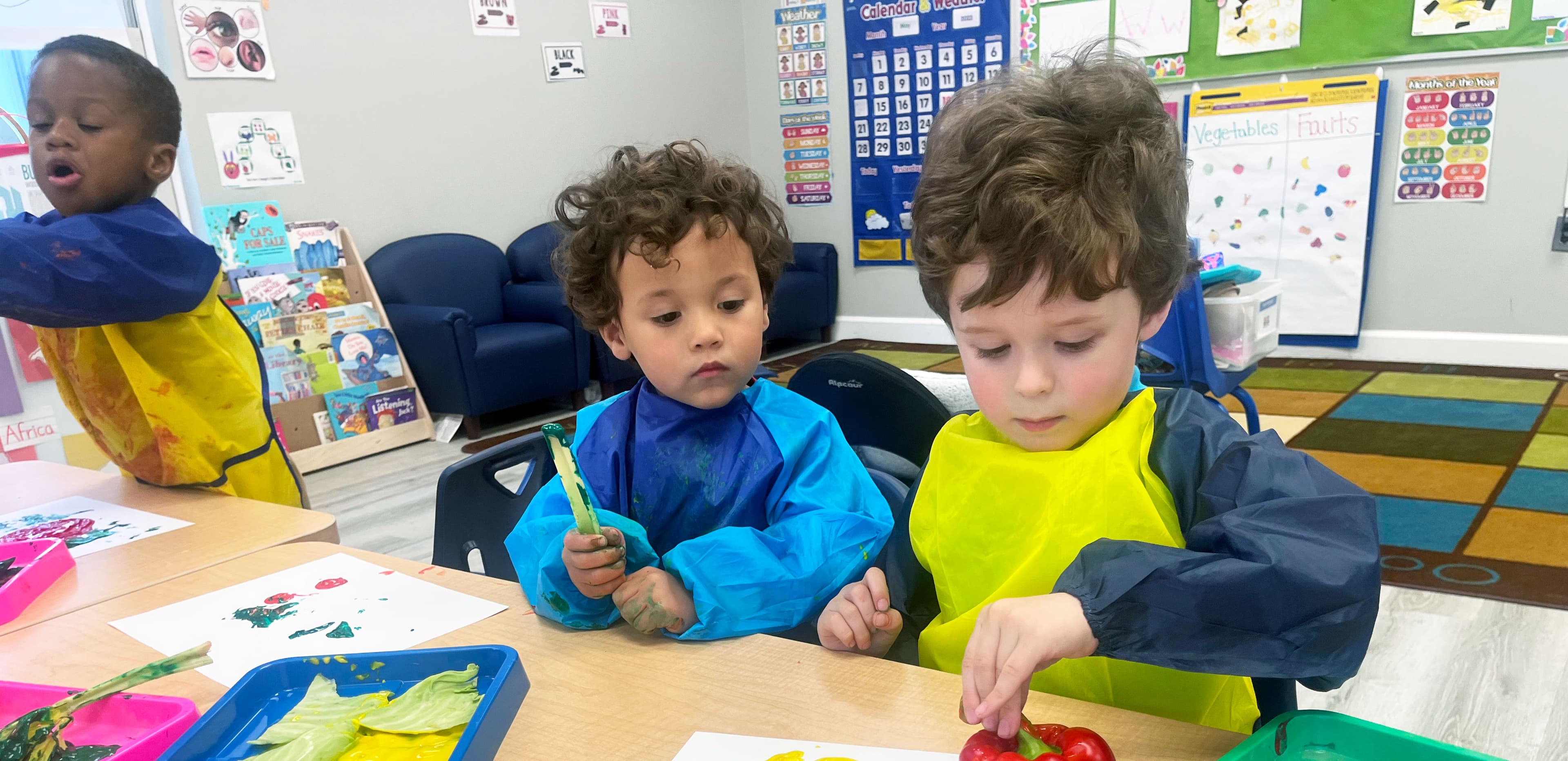 Two children in paint smocks making vegetable prints at a classroom table while a third child paints nearby