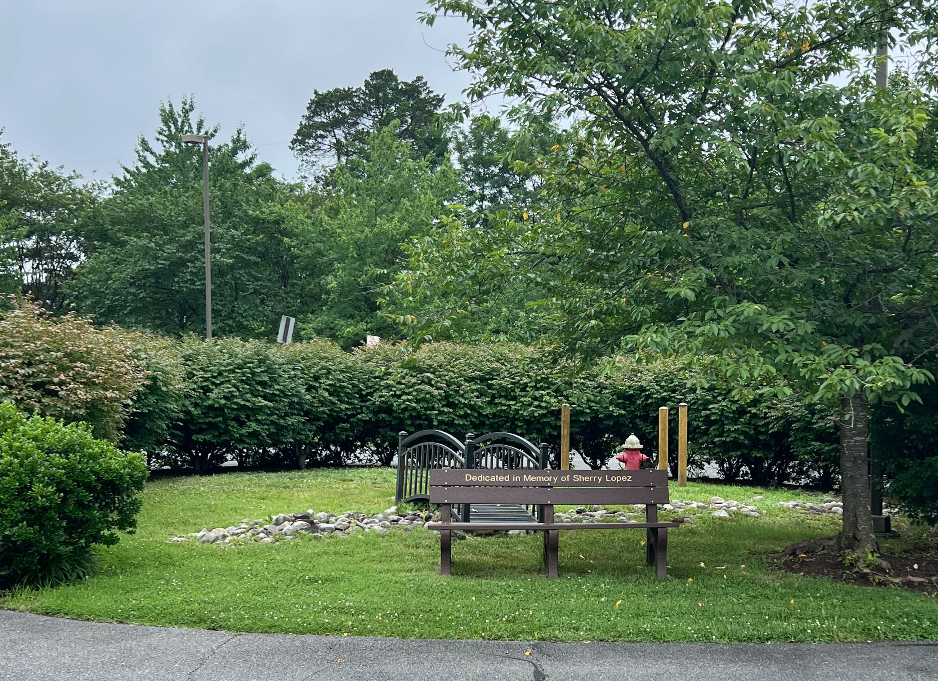 A memorial bench reading “Dedicated in Memory of Sherry Lopez” in a green garden with a small footbridge