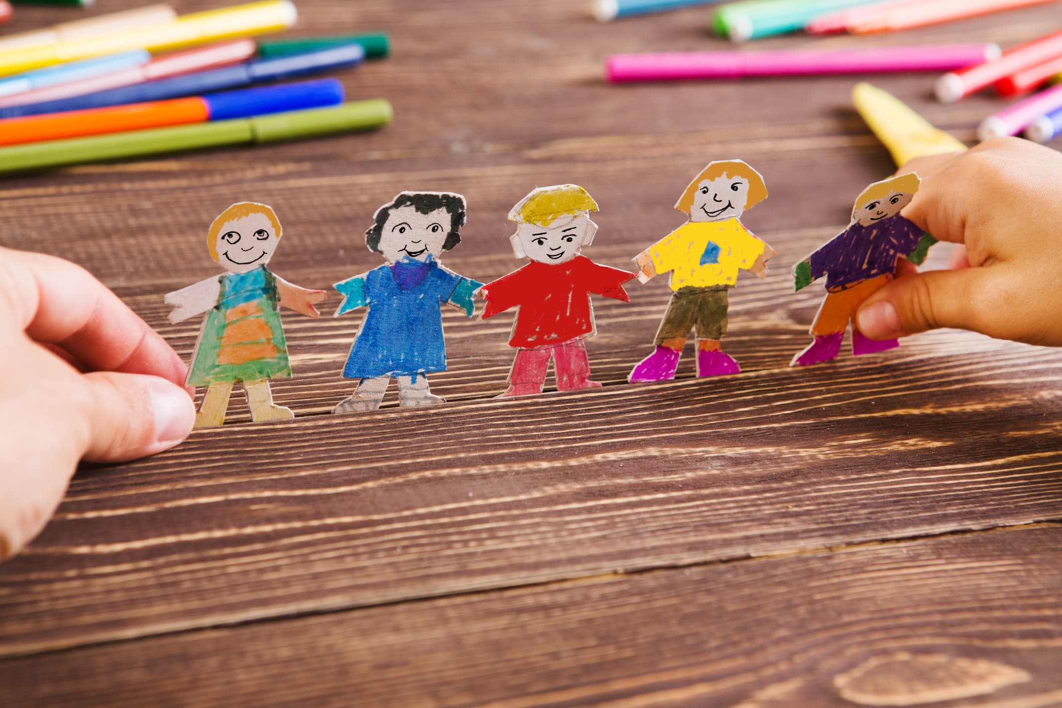 A chain of hand-drawn paper dolls holding hands on a wooden table surrounded by colored markers