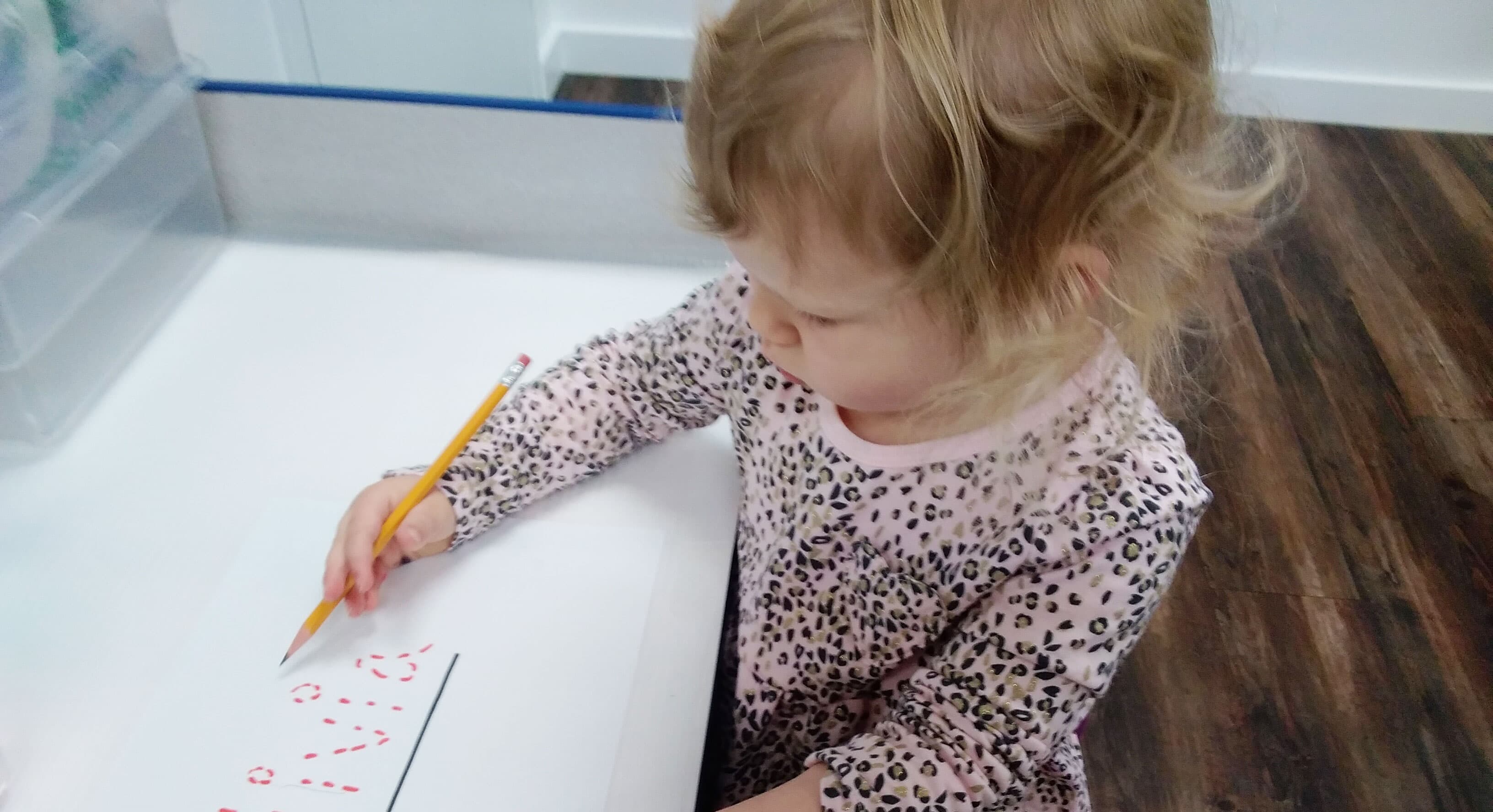 A toddler in a leopard-print top concentrating as she draws red marks on paper with a pencil