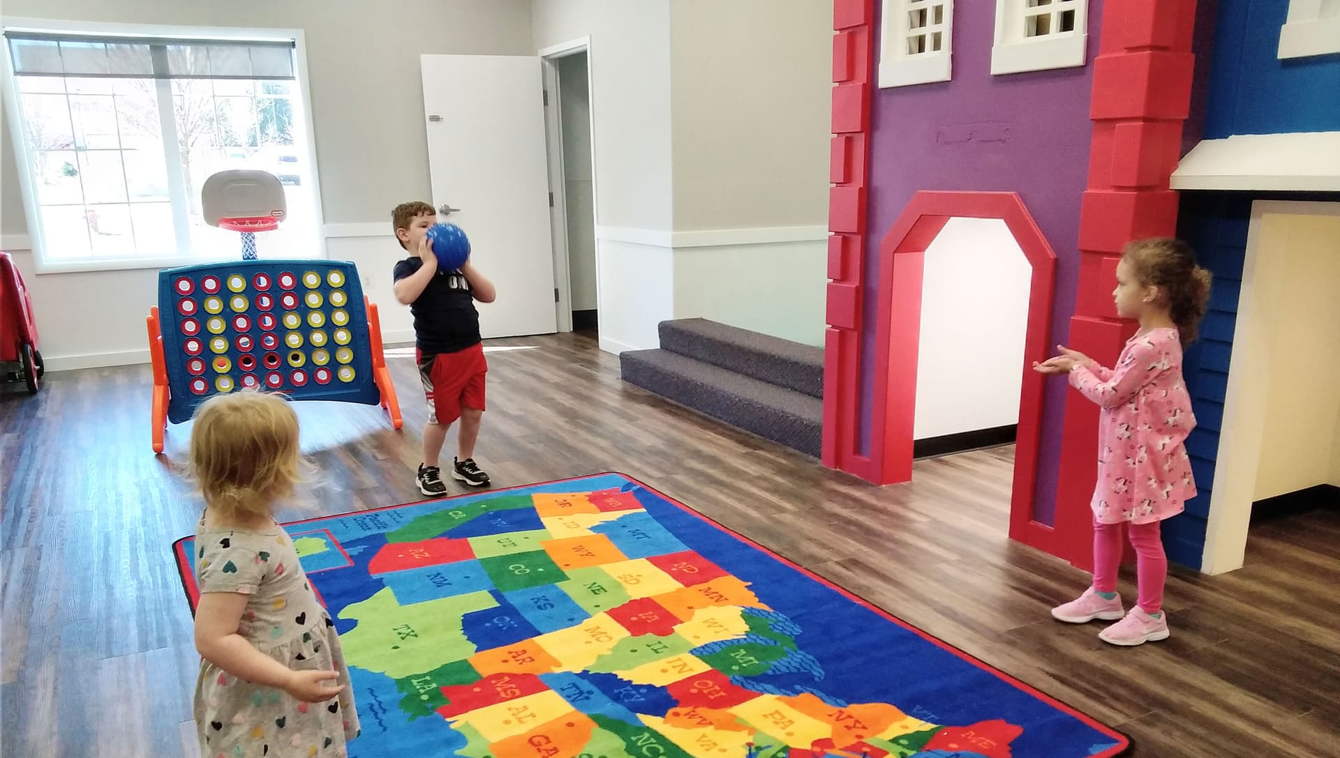 Children playing with a ball and a US map rug in the indoor playroom beside a red and purple playhouse