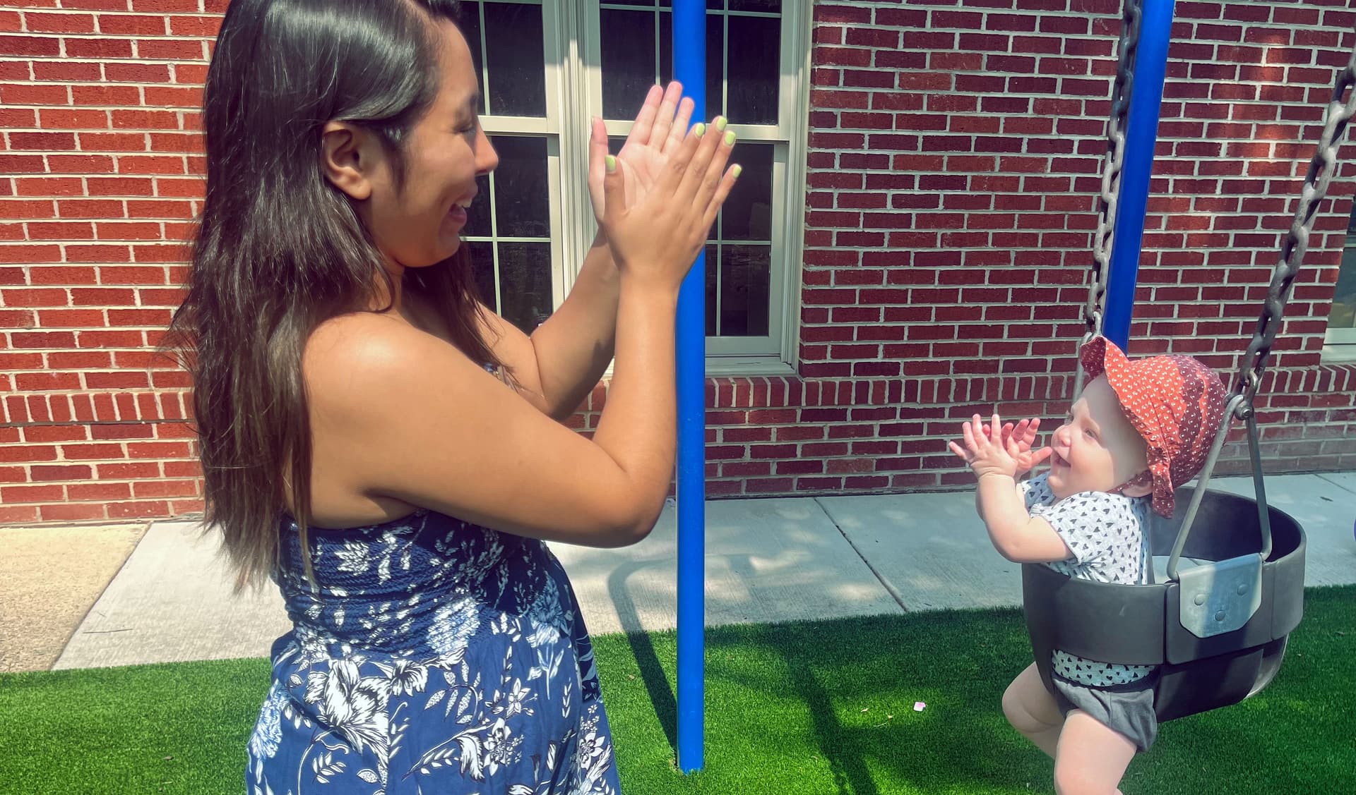 A teacher clapping hands with a smiling baby seated in a bucket swing on the playground